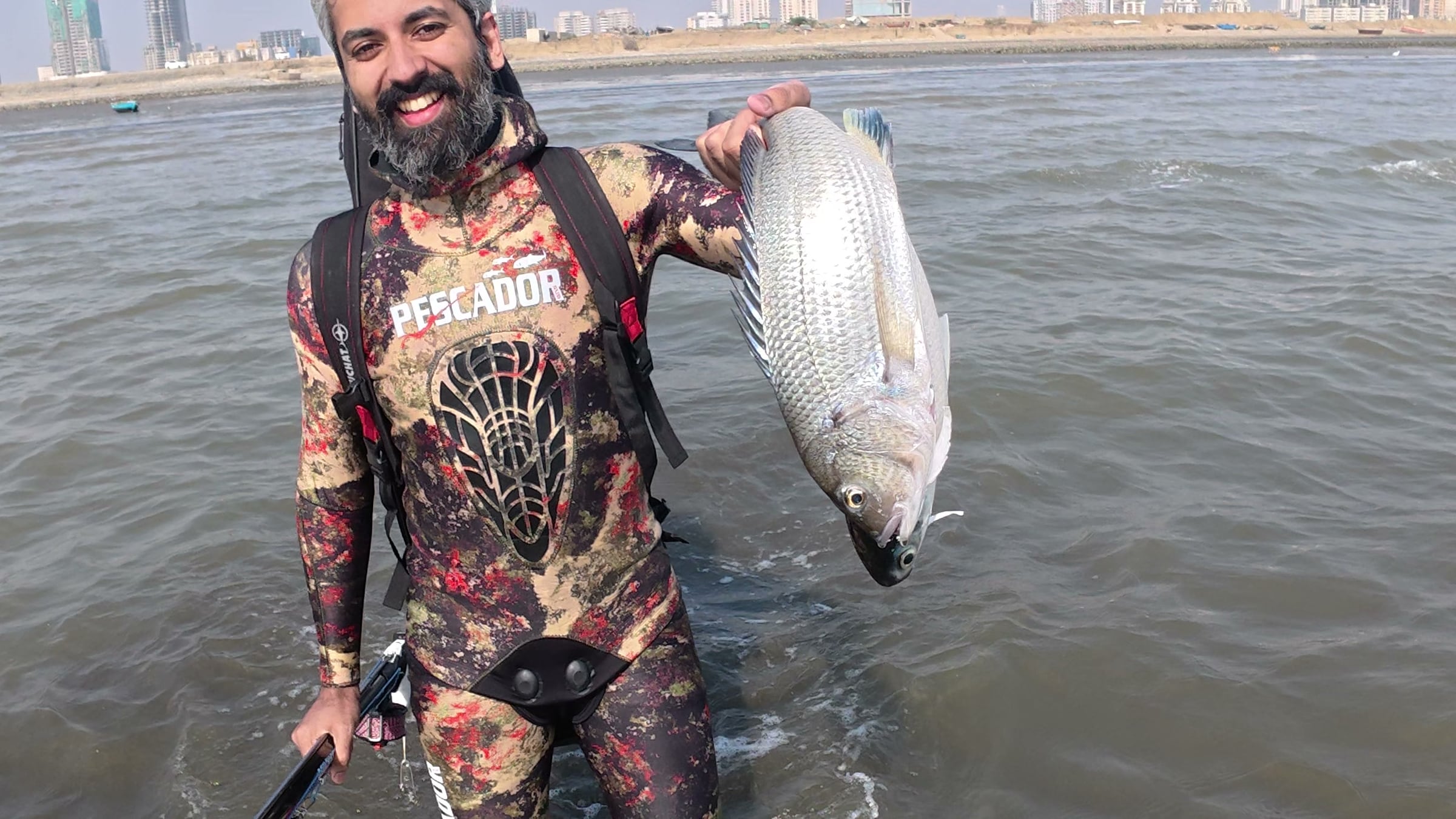Sameer wading out of the shallows in a camouflage wetsuit, holding a large silver fish, the Karachi shoreline low behind him