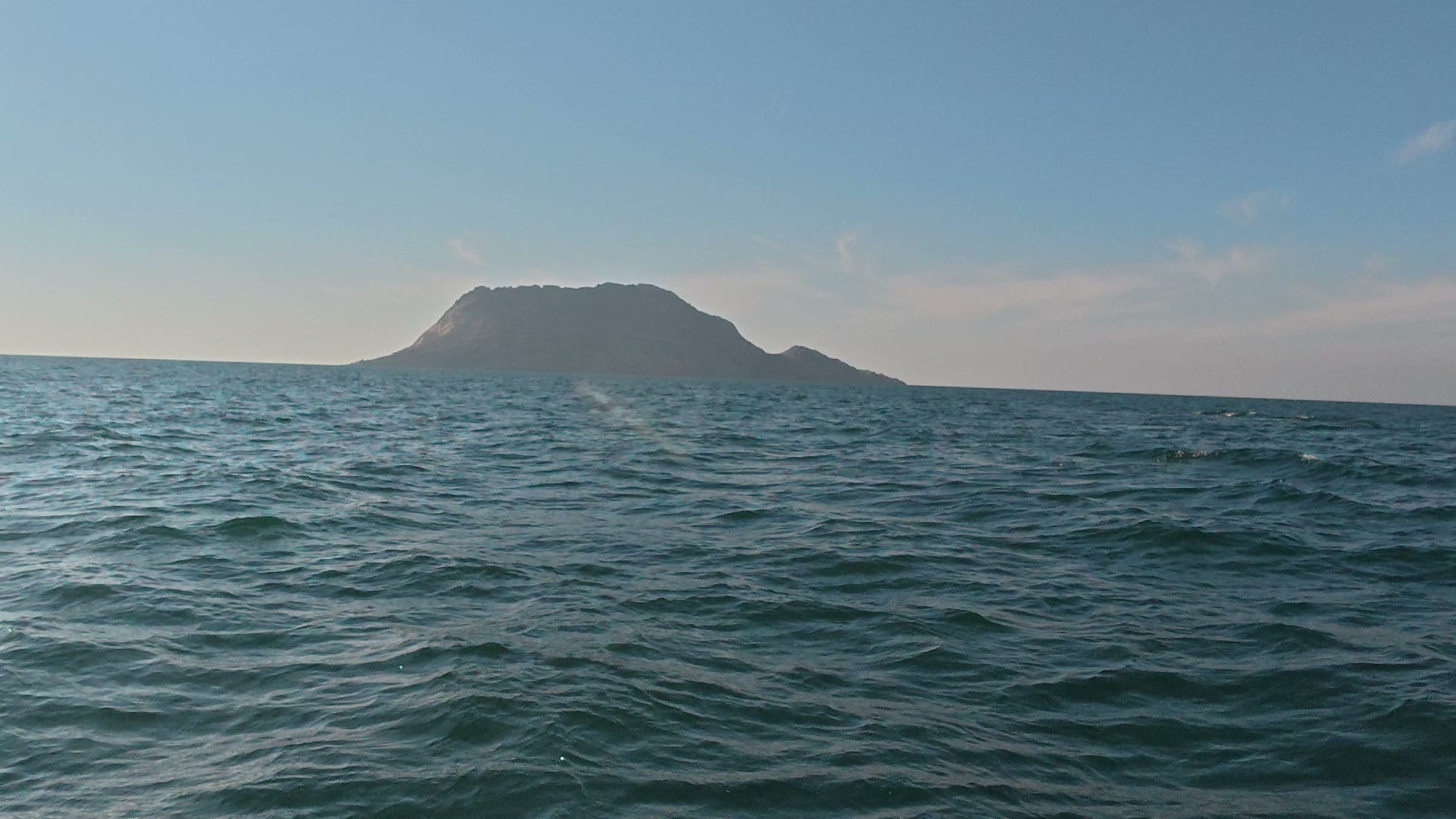 Charna Island as a low dark silhouette across flat open water, photographed from sea level on the way out to dive