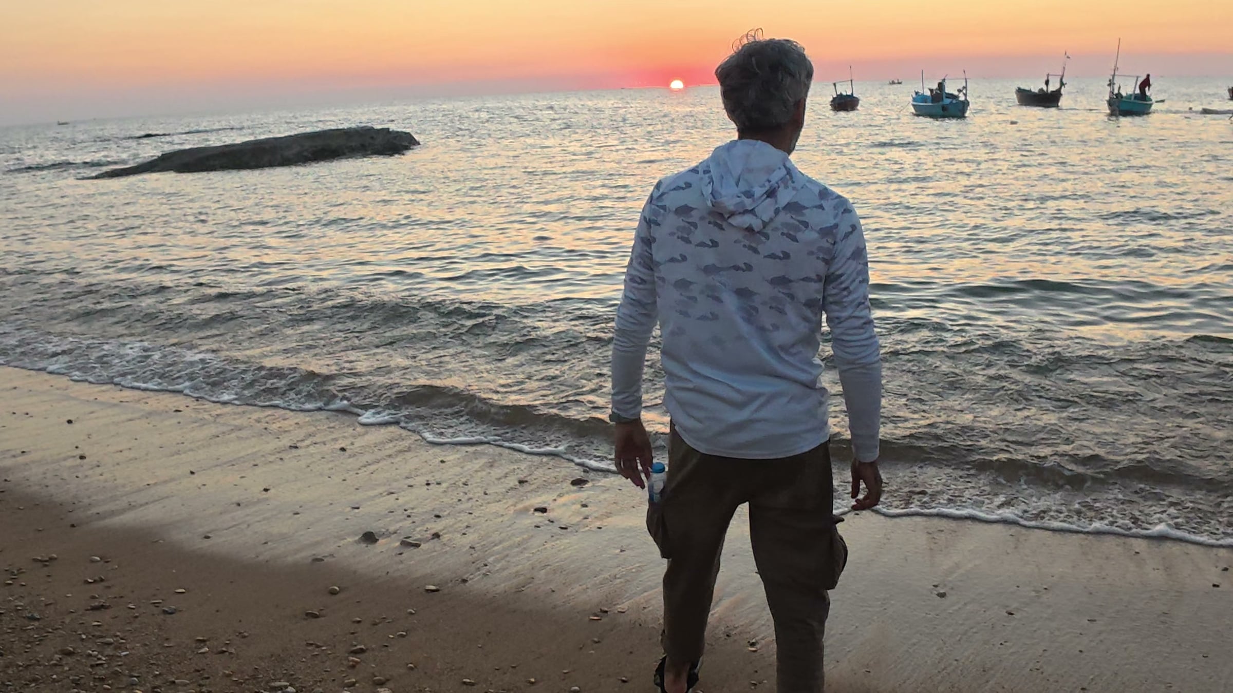 Sameer standing at the waterline at sunset with his back to the camera, wooden fishing boats moored just offshore