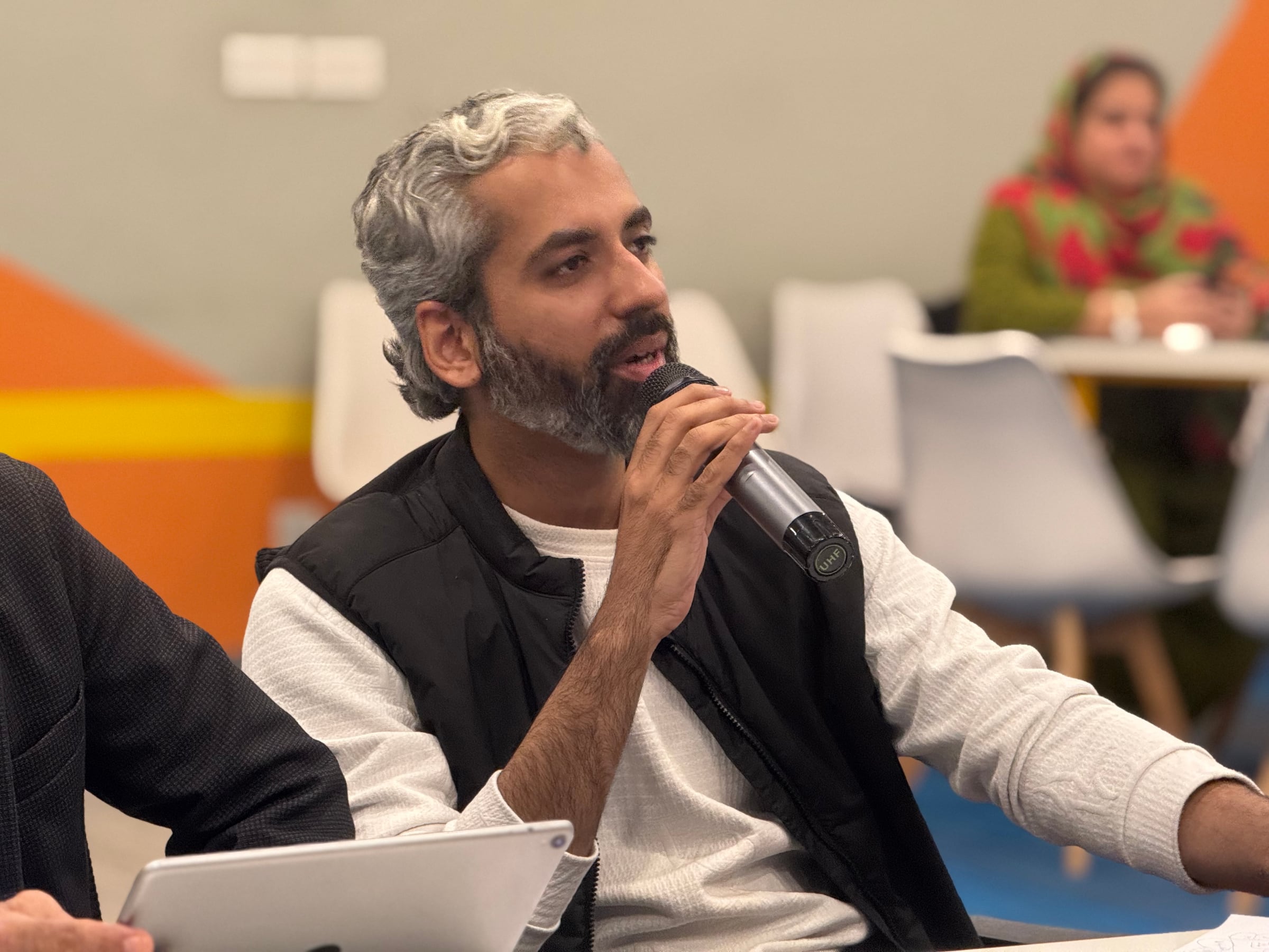 Sameer Ahmed Khan speaking into a handheld microphone during a panel discussion, orange seating behind him