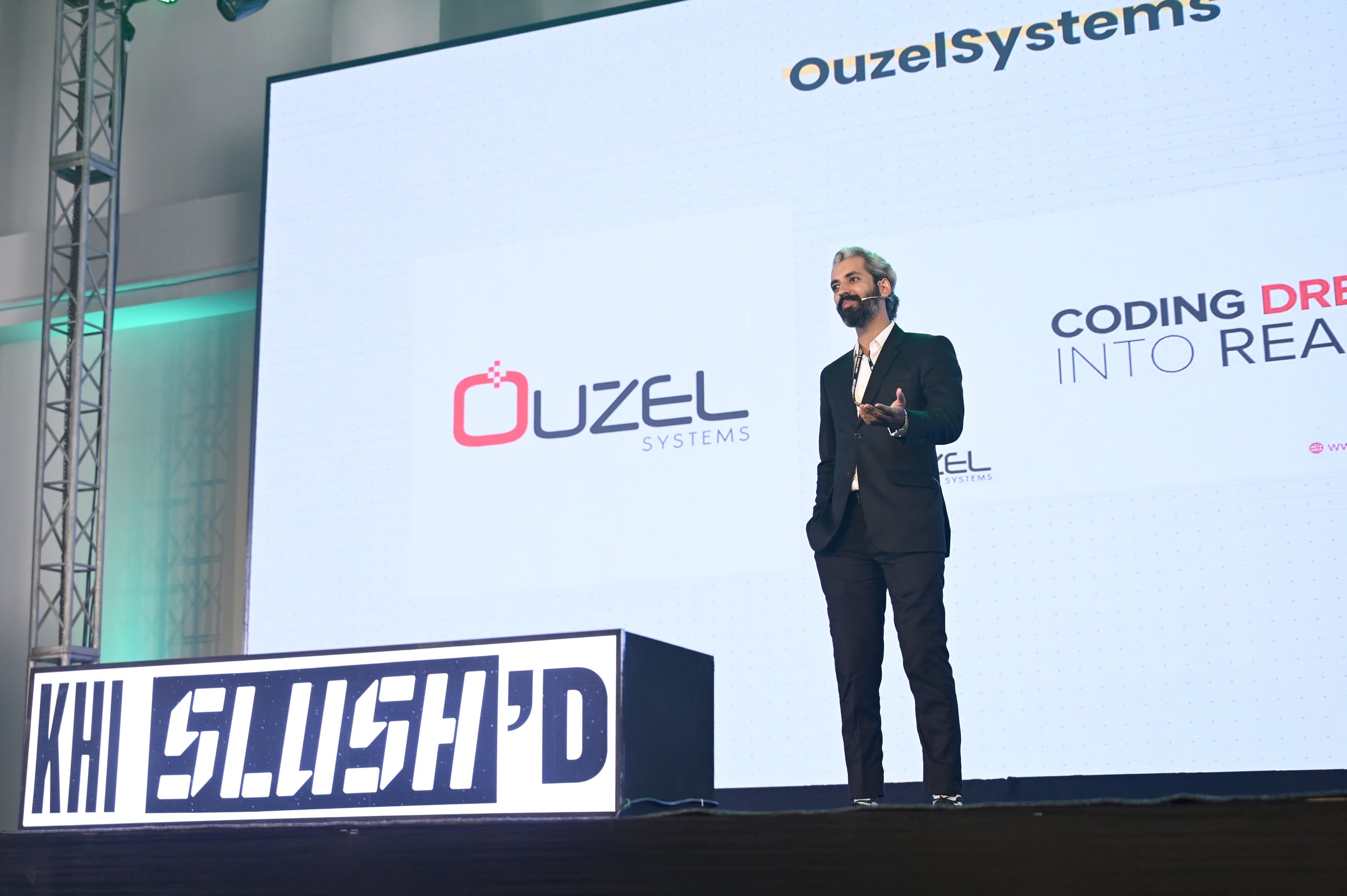 Sameer Ahmed Khan alone on a lit stage at Karachi Slush'D, a slide reading Coding Dreams Into Reality projected behind him